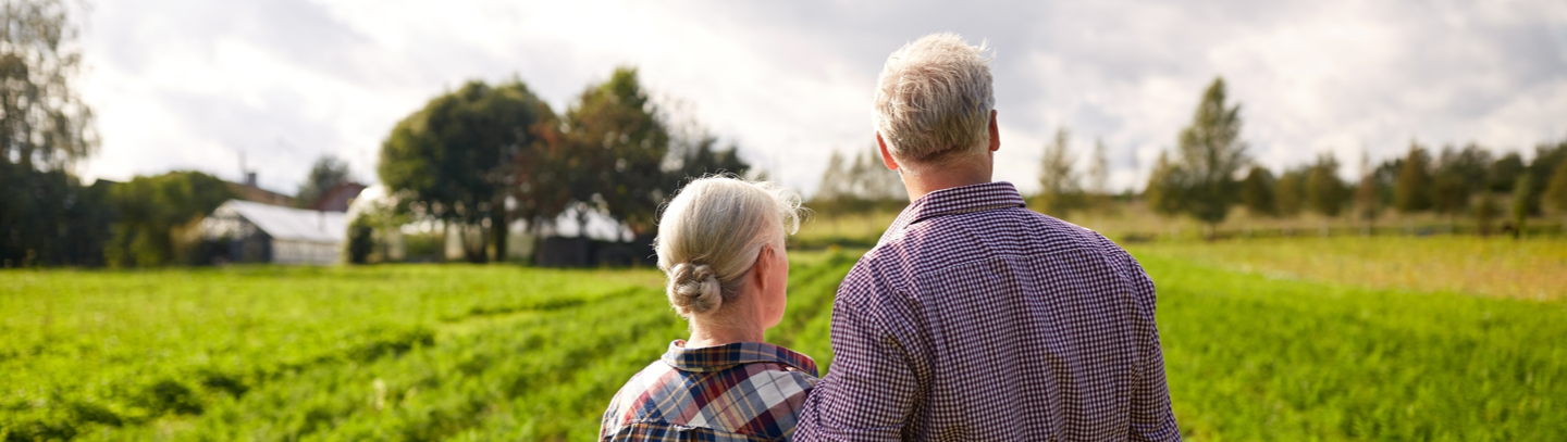 An older couple stands in their farmâ€™s field among the crops with their arms wrapped around each other's waists. 