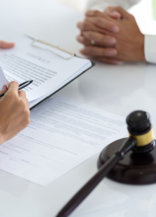 Two business people are sitting at a table, one holding paperwork with a gavel, and one with their hands folded in front of them. 