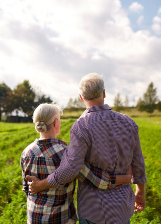 An older couple stands in their farmâ€™s field among the crops with their arms wrapped around each other's waists. 