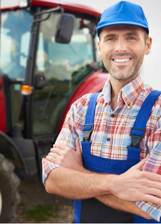 A male farmer stands outside in front of his red tractor parked in front of a barn. 