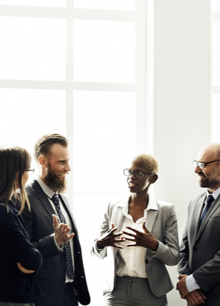 Nine professionally attired women and men stand in front of a large window wall discussing non-profit board insurance options.