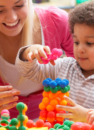 Female daycare instructor helps a male toddler sitting at table use round, colored plastic stacking shapes to build a tower. 