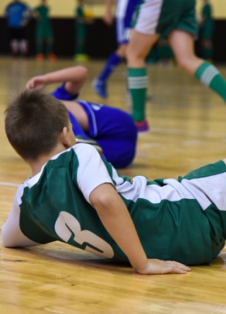 Two school-age soccer players fall down during a game in a gymnasium. 