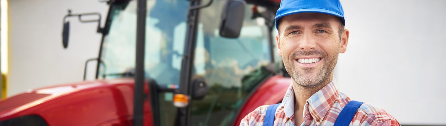 A male farmer stands outside in front of his red tractor parked in front of a barn. 