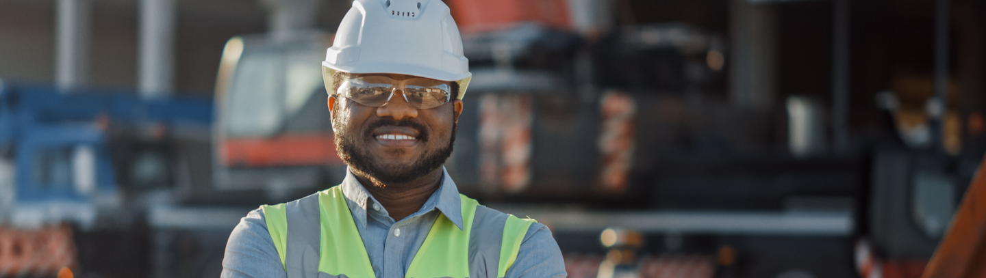 Smiling contractor wearing a safety hardhat, goggles, and vest stands outside a commercial construction site.