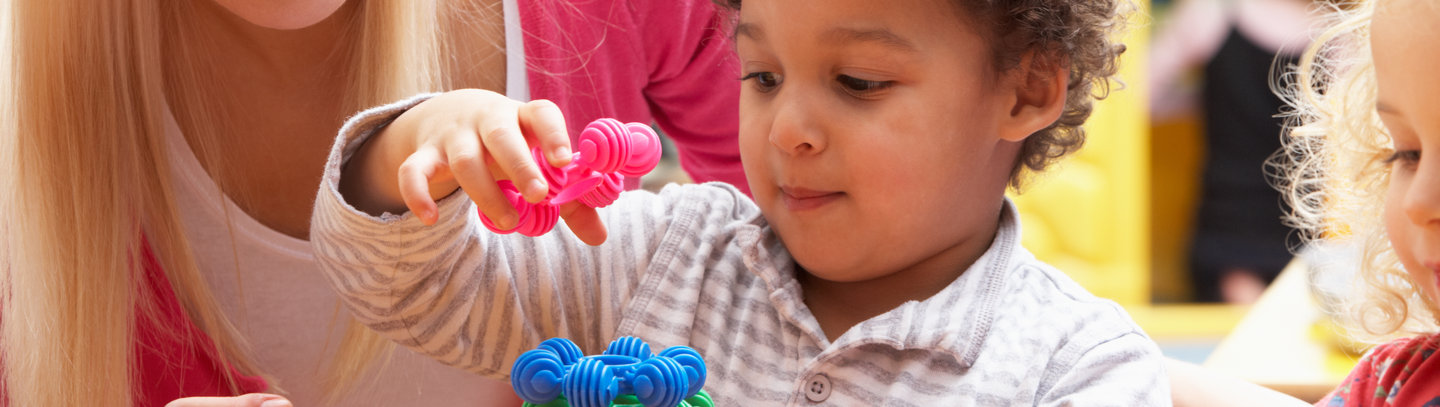 Female daycare instructor helps a male toddler sitting at table use round, colored plastic stacking shapes to build a tower. 