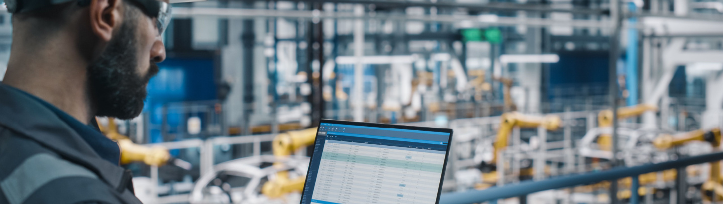 Worker looks at computer while standing in manufacturing plant. 