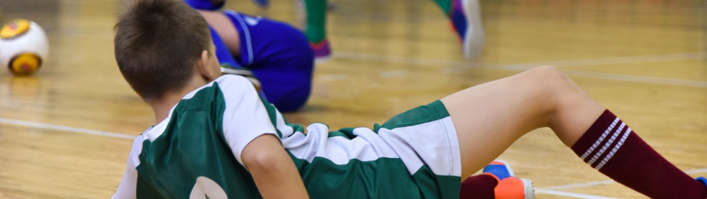 Two school-age soccer players fall down during a game in a gymnasium. 