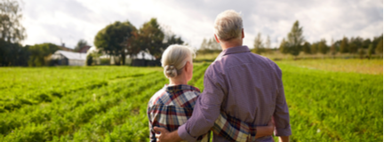 An older couple stands in their farm’s field among the crops with their arms wrapped around each other's waists. 