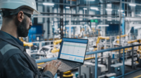 Worker looks at computer while standing in manufacturing plant. 