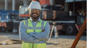 Smiling contractor wearing a safety hardhat, goggles, and vest stands outside a commercial construction site.