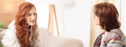 A woman sits on a couch in a mental health organization, talking with a therapist in a chair who holds a tablet computer.