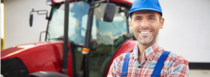 A male farmer stands outside in front of his red tractor parked in front of a barn. 