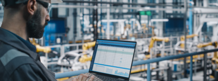 Worker looks at computer while standing in manufacturing plant. 