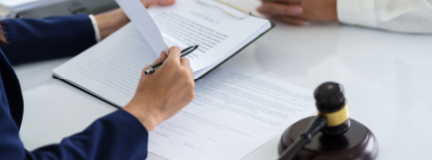 Two business people are sitting at a table, one holding paperwork with a gavel, and one with their hands folded in front of them. 