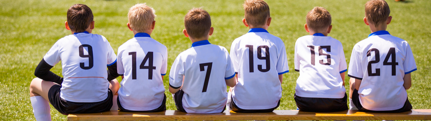 Six young boys seated on bench at soccer game with backs to camera wearing numbered white jerseys.
