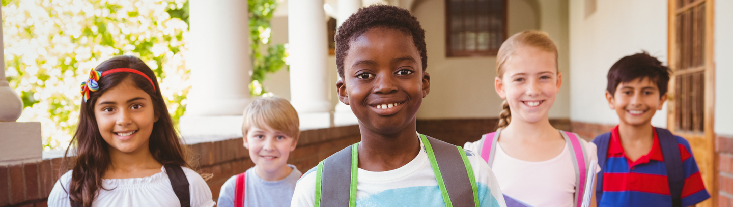 Group of four elementary age school children wearing backpacks in outdoor corridor, looking at camera.   