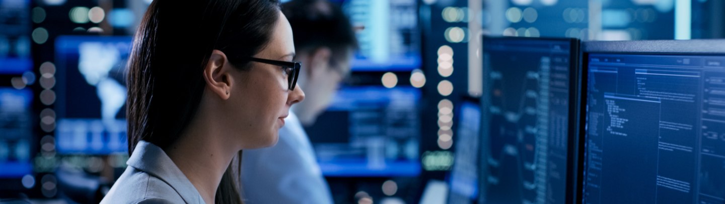 Female engineer watches monitor screens while in the background a person is working and monitors show various information.