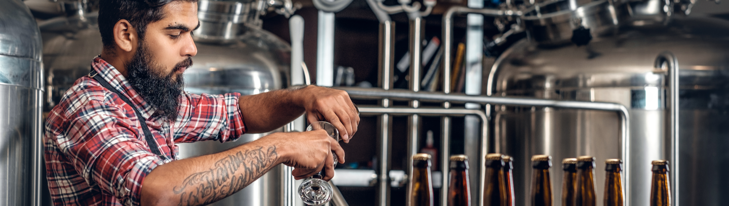 Bearded brewmaster pours samples of craft beer with beer tanks in the background. 