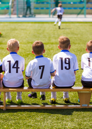 Six young boys seated on bench at soccer game with backs to camera wearing numbered white jerseys.