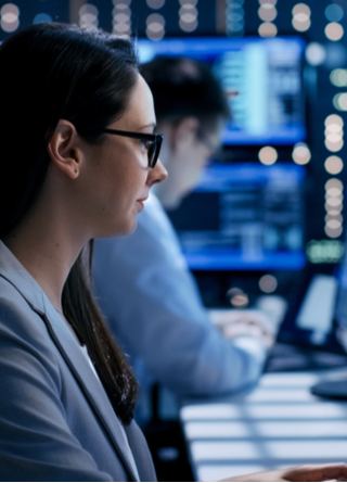 Female engineer watches monitor screens while in the background a person is working and monitors show various information.