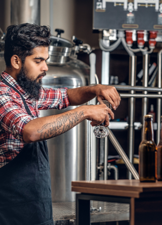 Bearded brewmaster pours samples of craft beer with beer tanks in the background. 