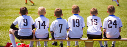 Six young boys seated on bench at soccer game with backs to camera wearing numbered white jerseys.