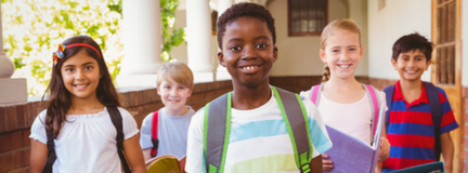 Group of four elementary age school children wearing backpacks in outdoor corridor, looking at camera.   