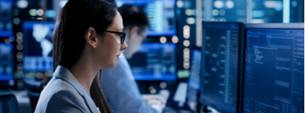 Female engineer watches monitor screens while in the background a person is working and monitors show various information.