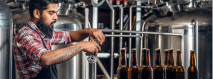 Bearded brewmaster pours samples of craft beer with beer tanks in the background. 